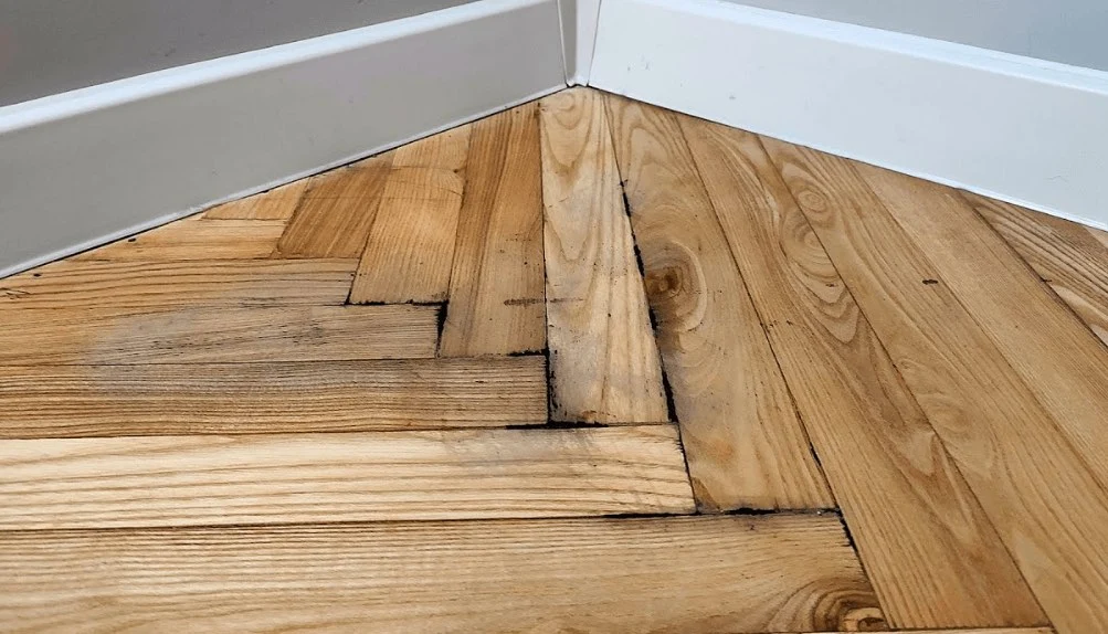 Dark water staining along the seams of a hardwood floor beside the baseboard